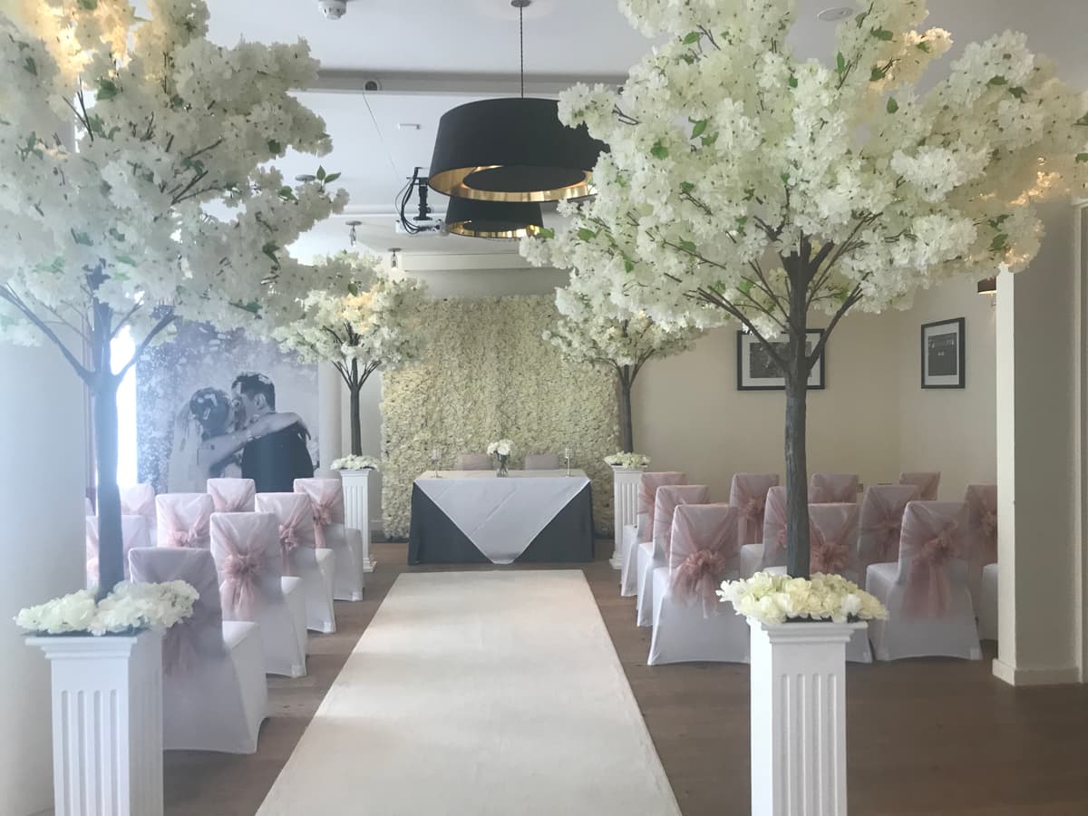 Civil ceremony room with white blossom trees, pink sash chairs and a floral wall backdrop.