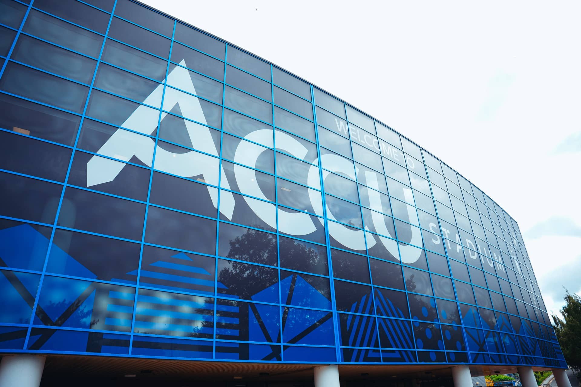 Exterior glass facade of Accu Stadium in Huddersfield with large blue branding signage.