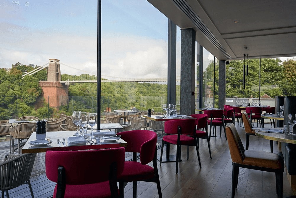 Restaurant interior with pink chairs, laid tables and views of Clifton Suspension Bridge.