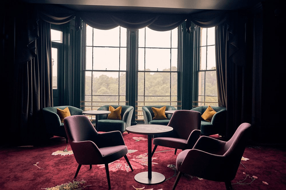 Lounge seating area with velvet armchairs, round tables and large bay windows overlooking trees.