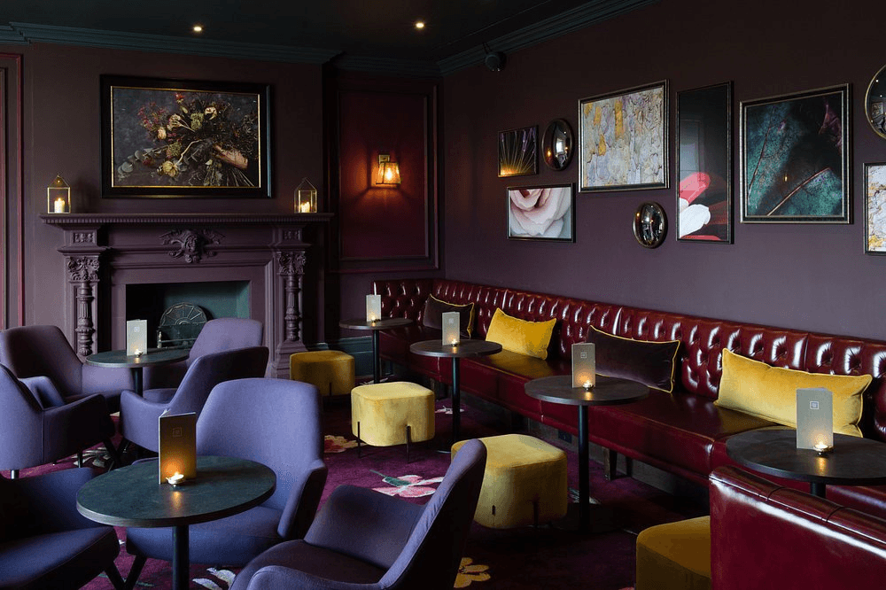 Dimly lit bar lounge with red leather banquettes, purple armchairs and candlelit tables.