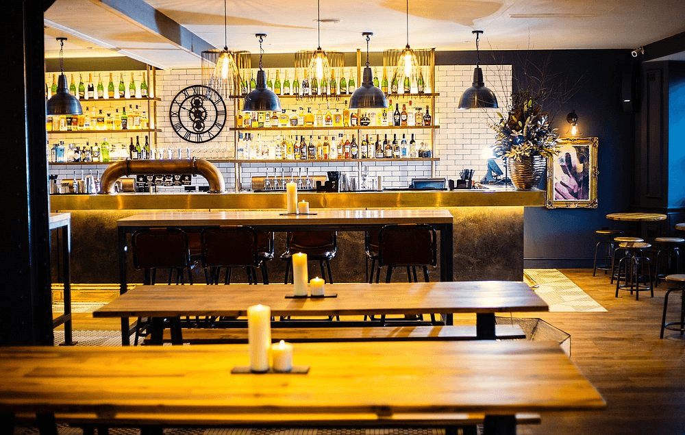 Candlelit wooden tables in front of a well-stocked bar with industrial pendant lights.