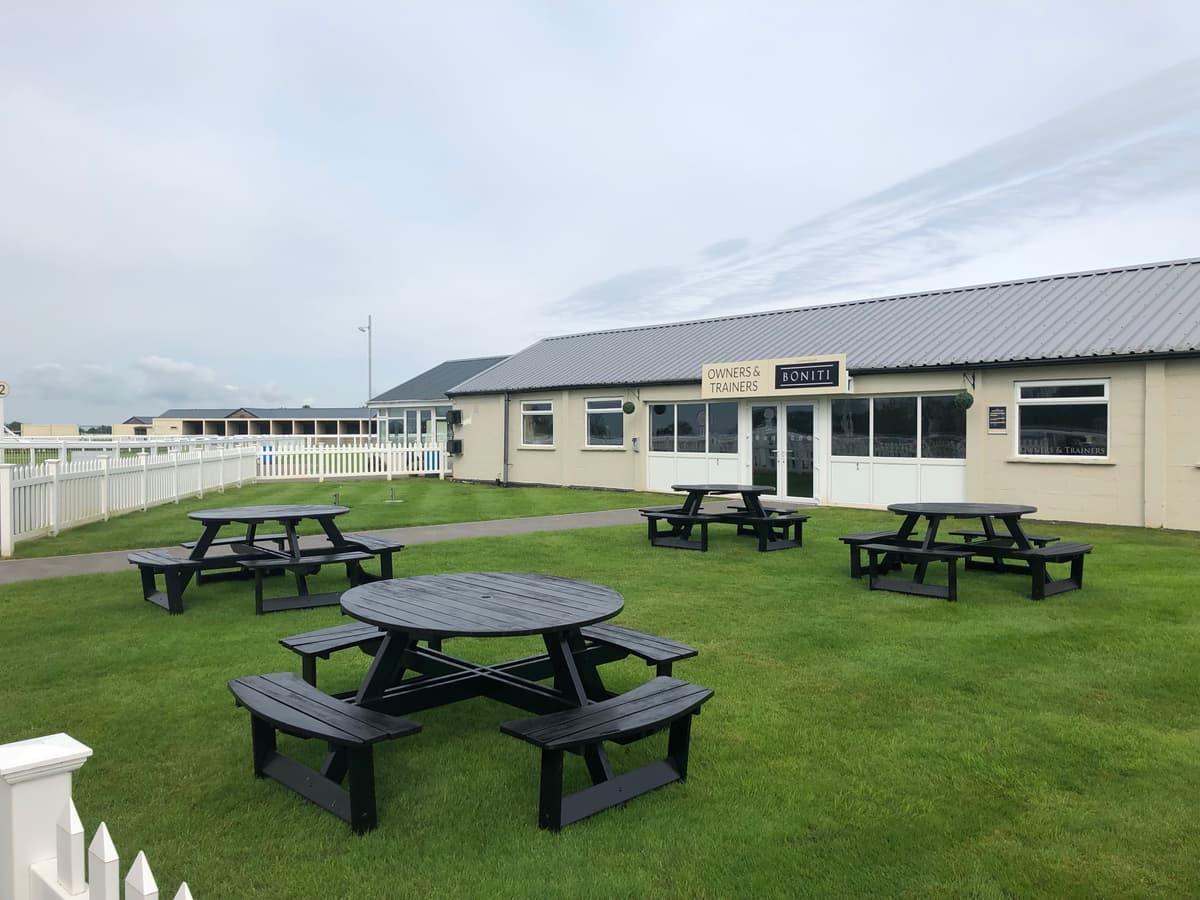 Outdoor seating area with black picnic tables on a lawn at Bath Racecourse.