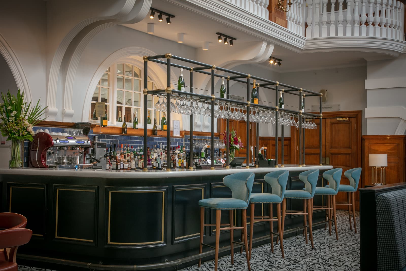The hotel bar at Chilworth Manor with teal stools, overhead glass rack and ornate white arches.