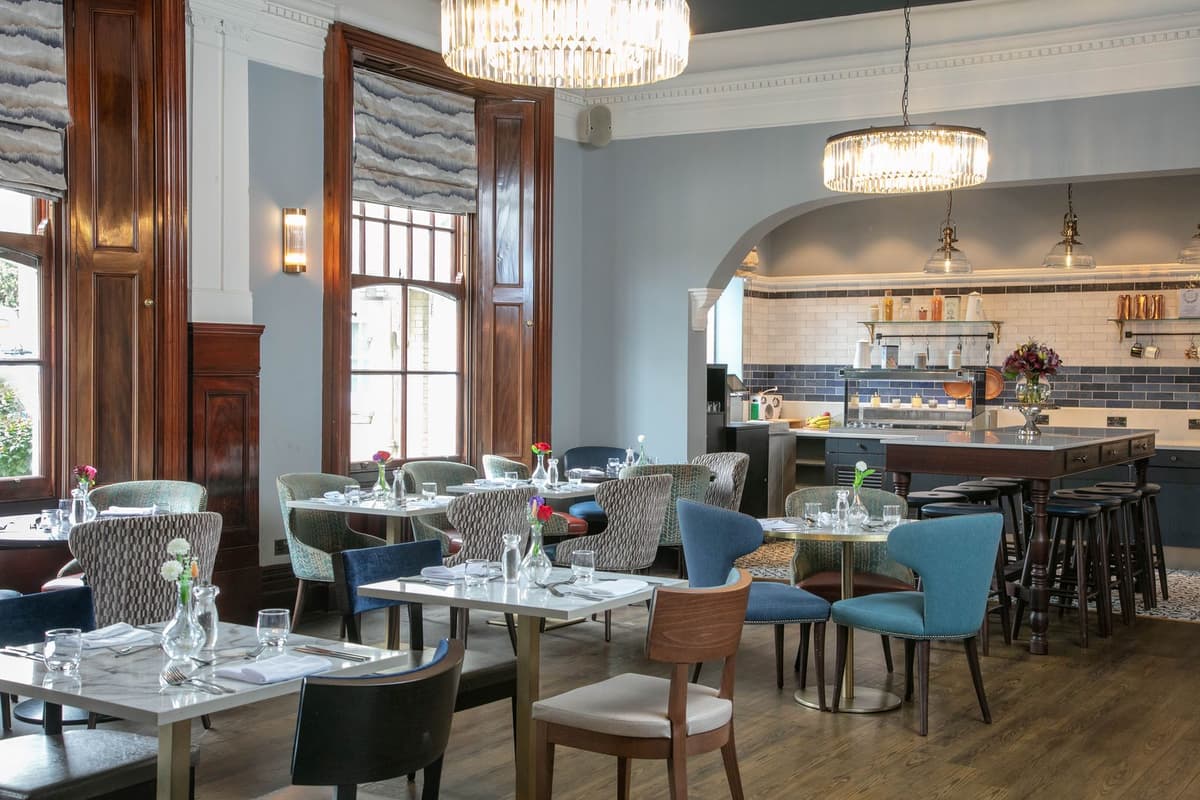 Bright restaurant interior with marble tables, mixed chairs and an open kitchen beyond.