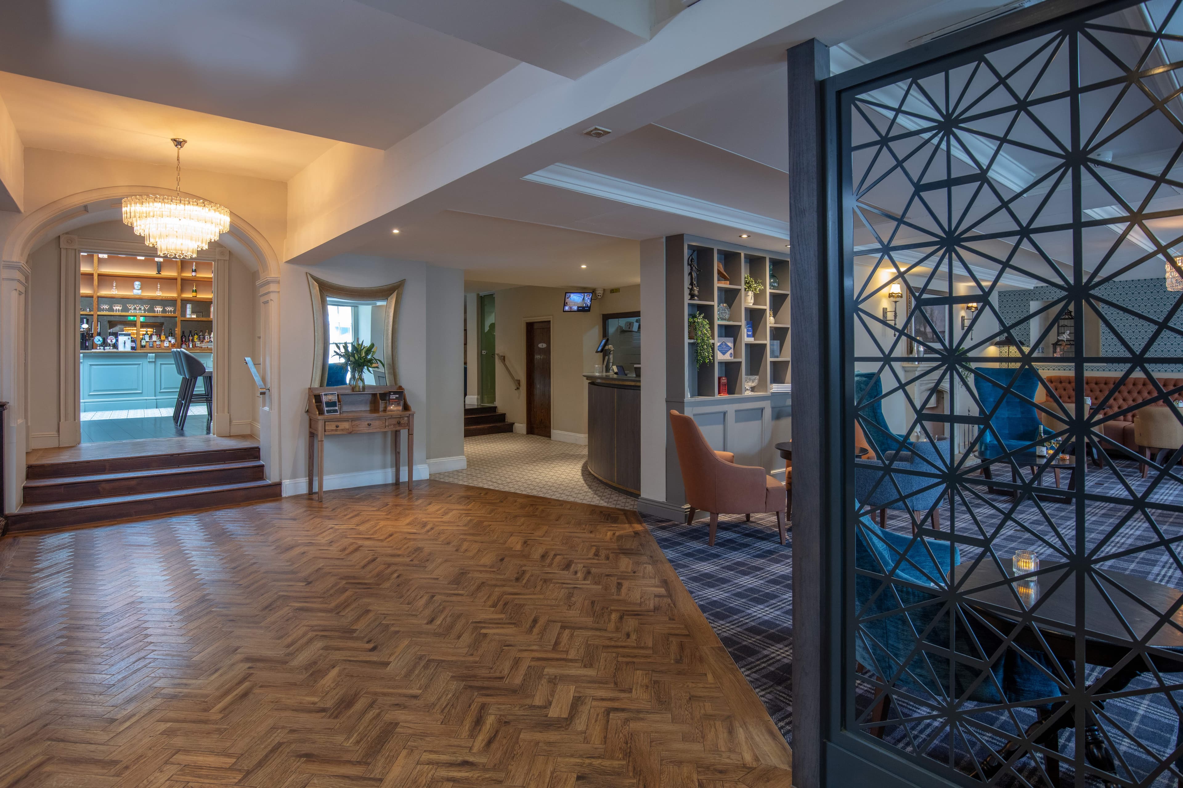 Hotel reception with parquet flooring, chandelier, arched bar entrance and a blue lounge beyond a decorative screen