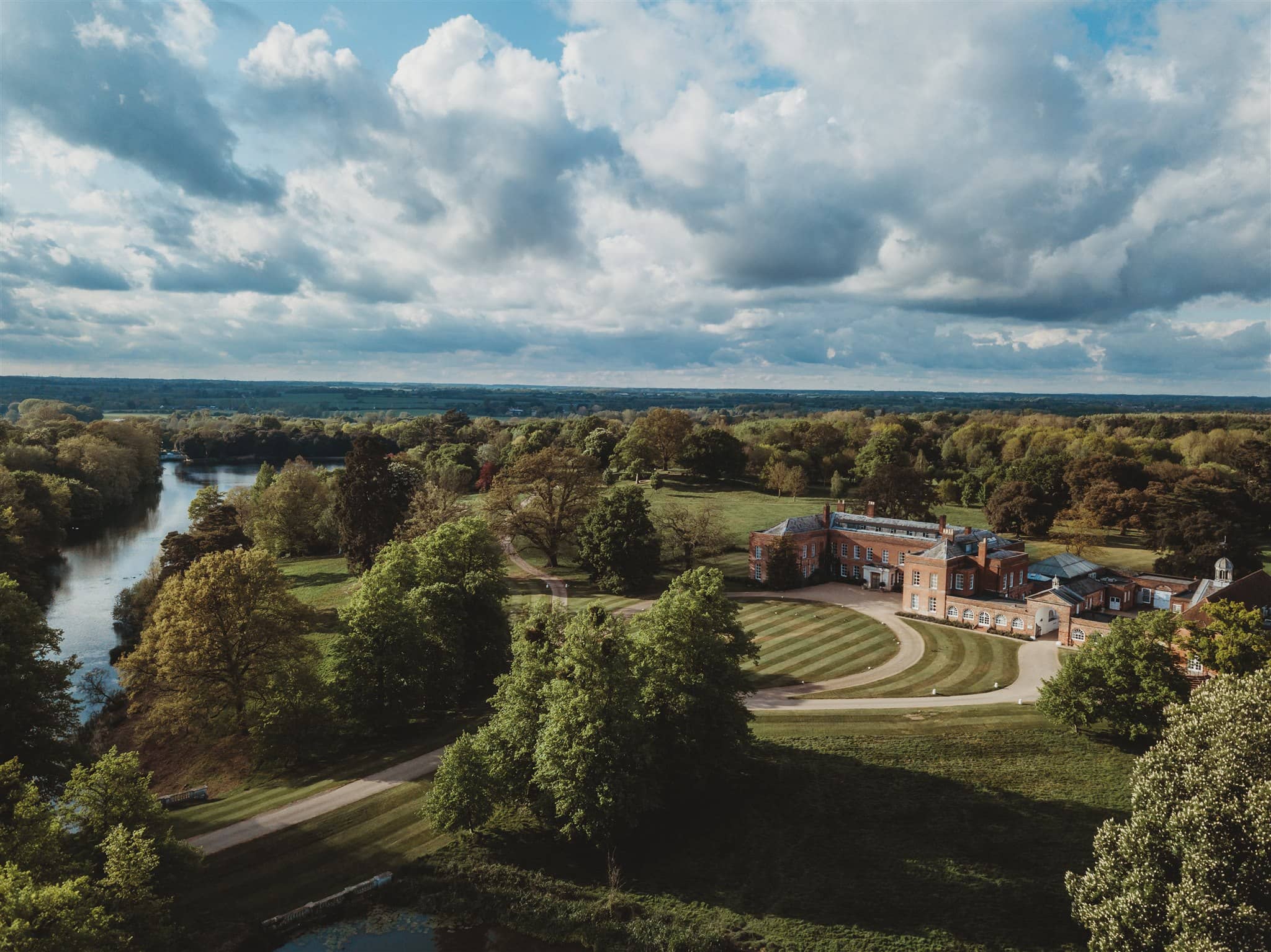 Aerial view of Braxted Park Estate's red brick manor house and landscaped grounds beside a river.