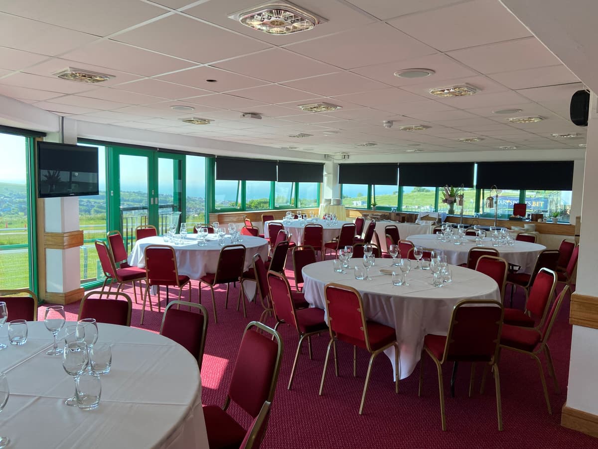 Bright function room at Brighton Racecourse with round tables, red chairs and panoramic views.