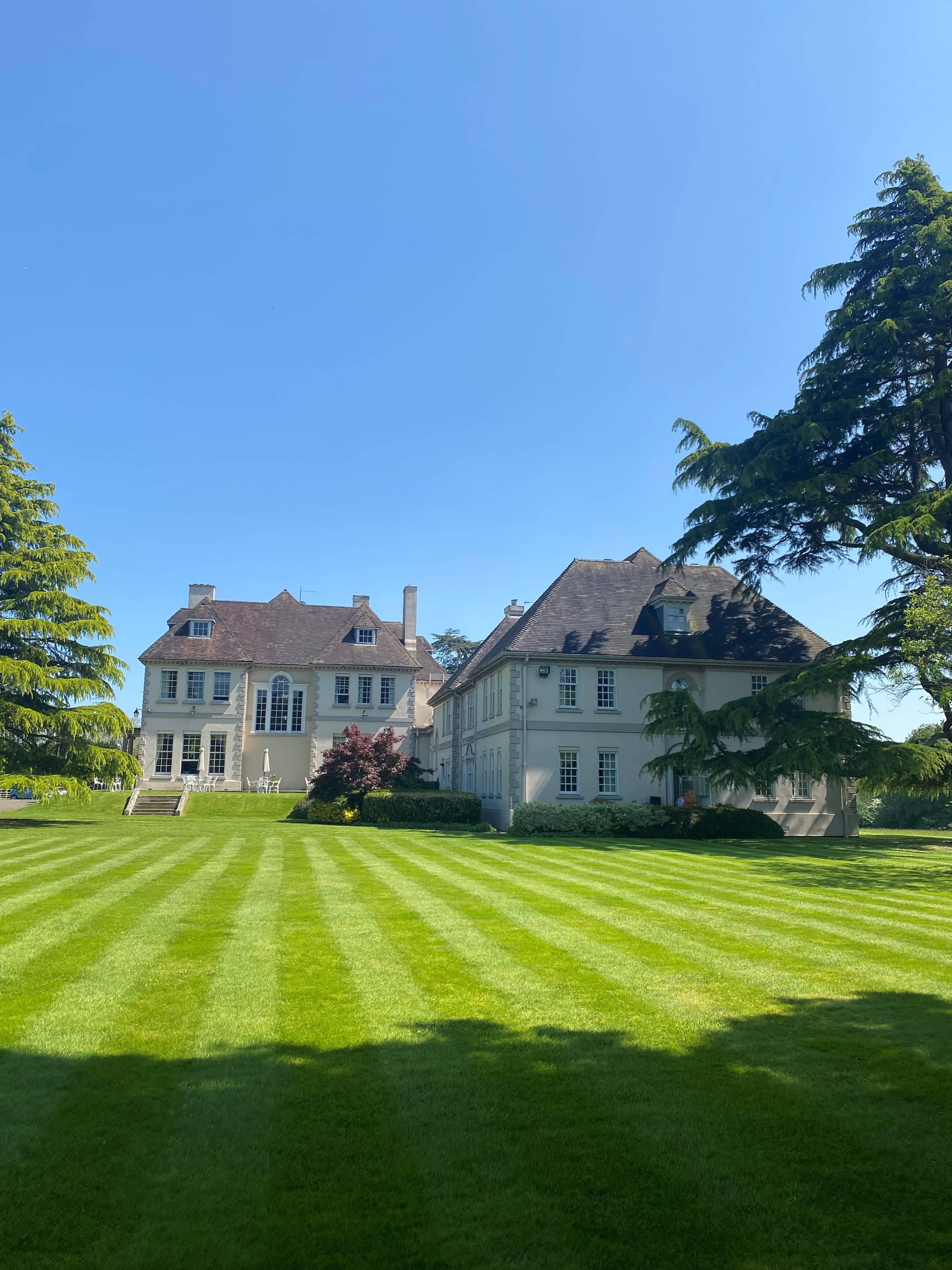Brockencote Hall exterior with striped lawns, mature cedar trees and clear blue sky.