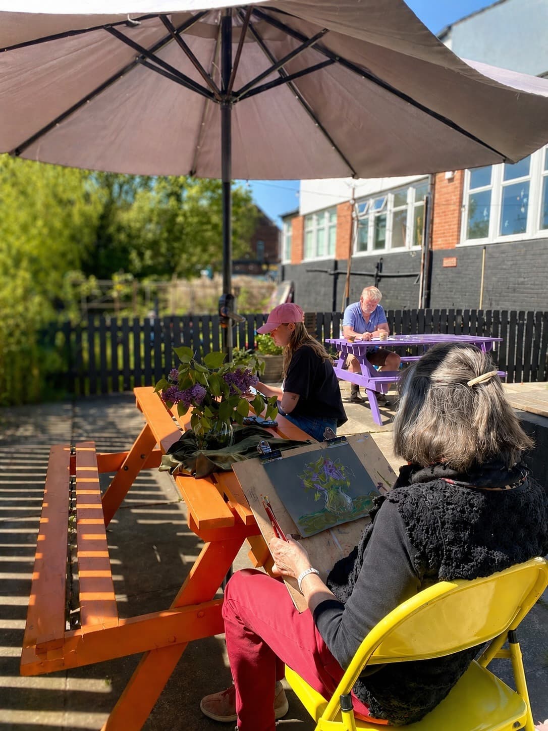 People drawing and painting outdoors at brightly coloured picnic tables in a sunny garden.
