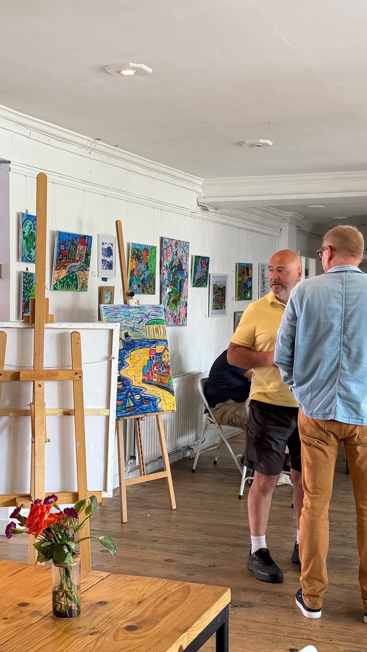 Two men talking beside colourful paintings on easels and walls in a bright gallery room.