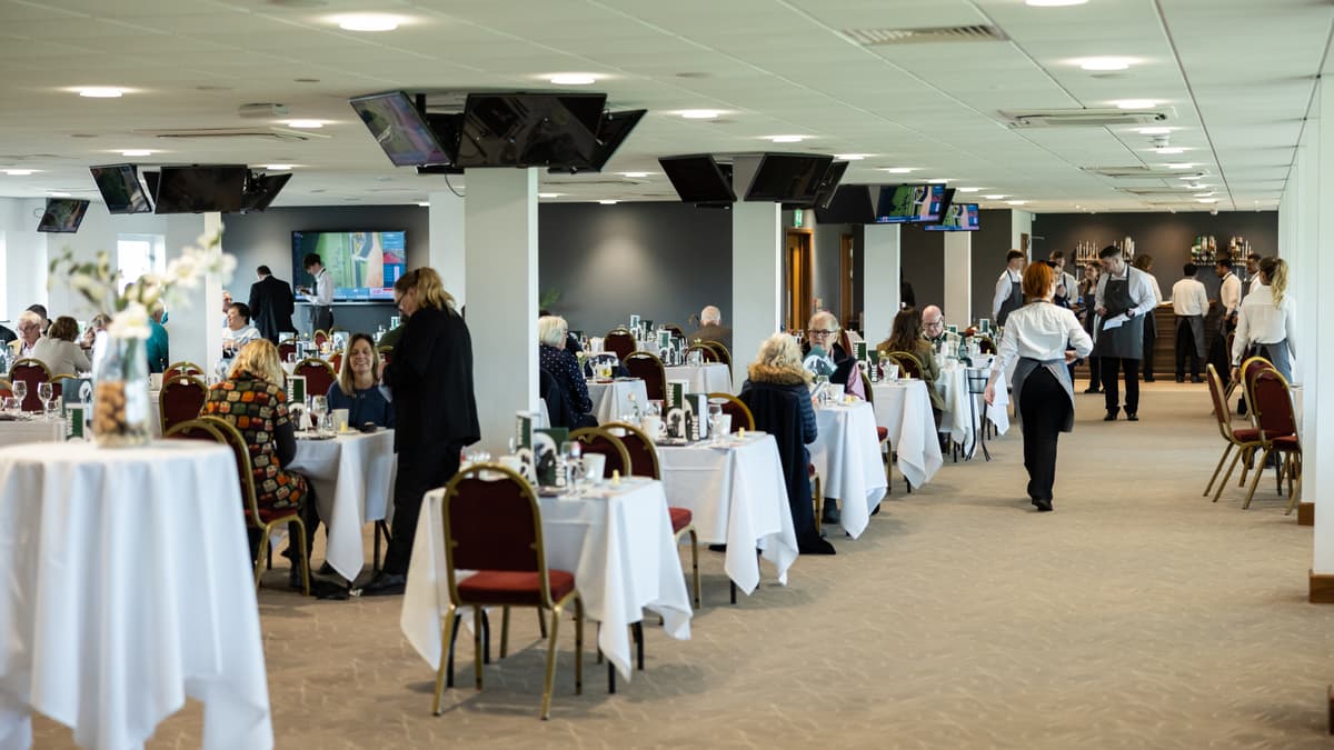Guests dining at white-clothed tables in a spacious hospitality suite with ceiling-mounted screens.