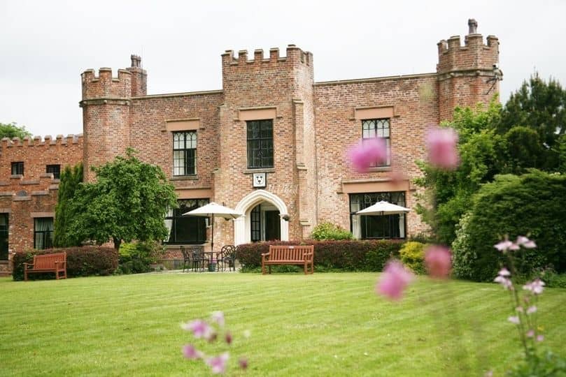 The red brick castellated exterior of Crabwall Manor with lawns and garden flowers.