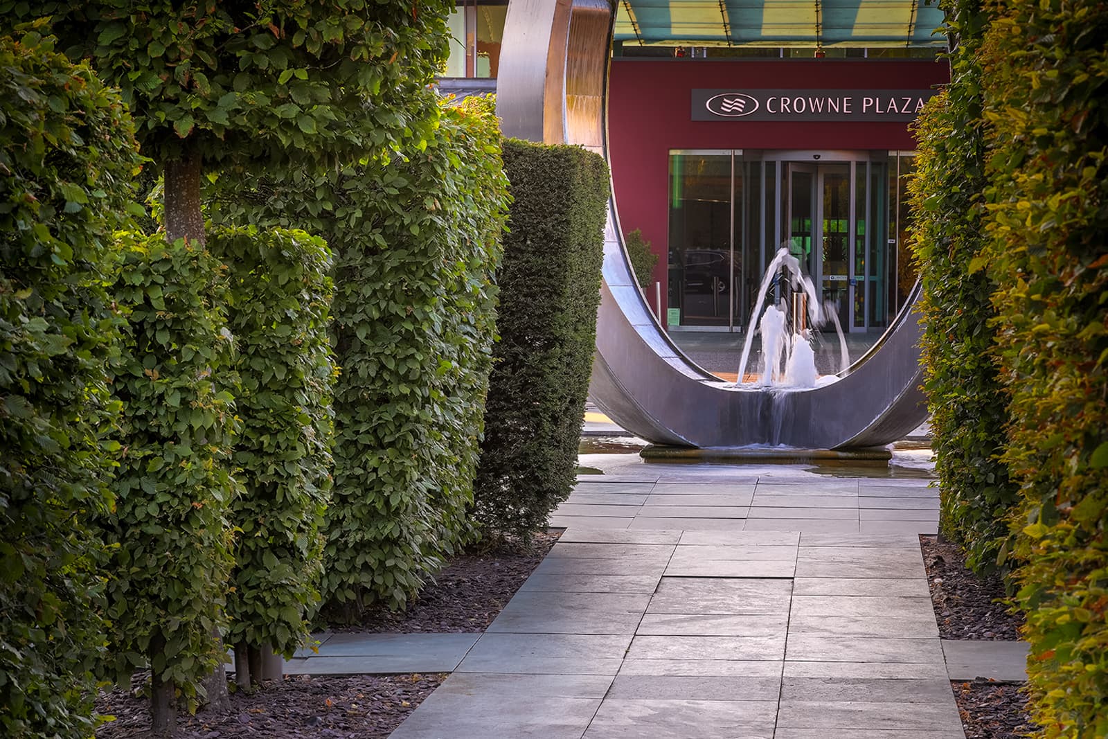 Stone pathway leading to the Crowne Plaza Marlow entrance flanked by hedgerows and a fountain sculpture.