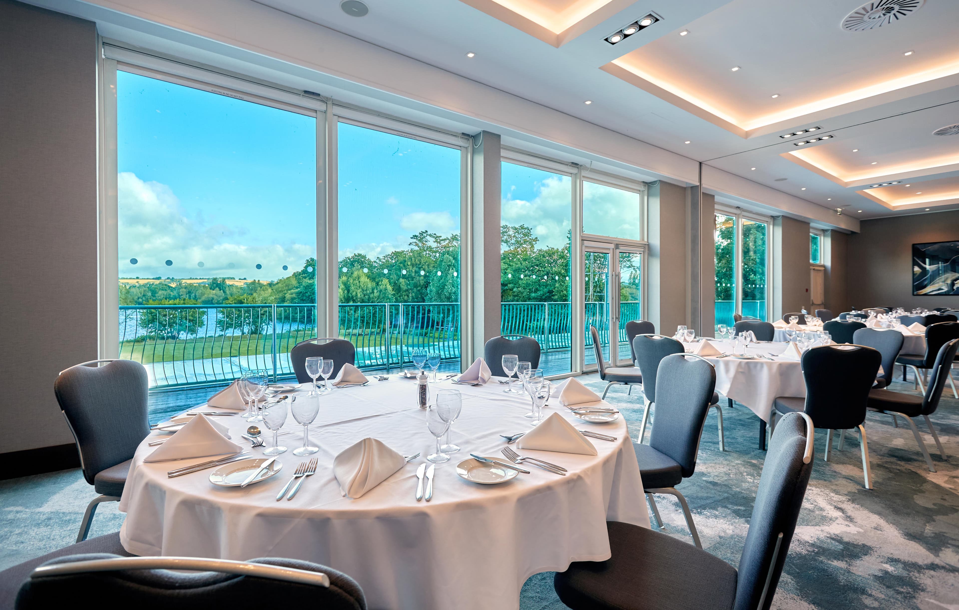 Banqueting room with round tables set for dining and views over countryside and lake.