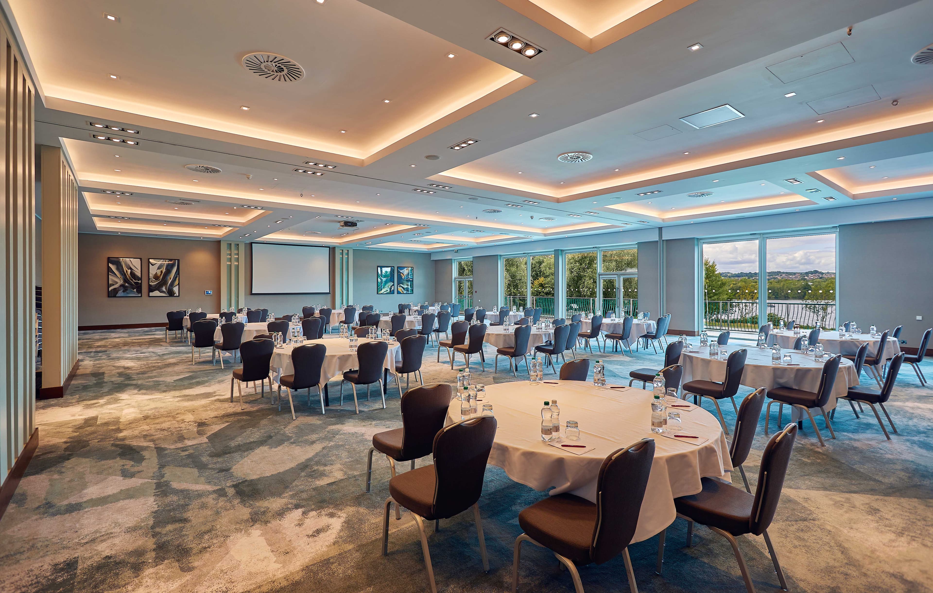 Spacious conference room with round tables, projection screen and floor-to-ceiling windows overlooking trees.