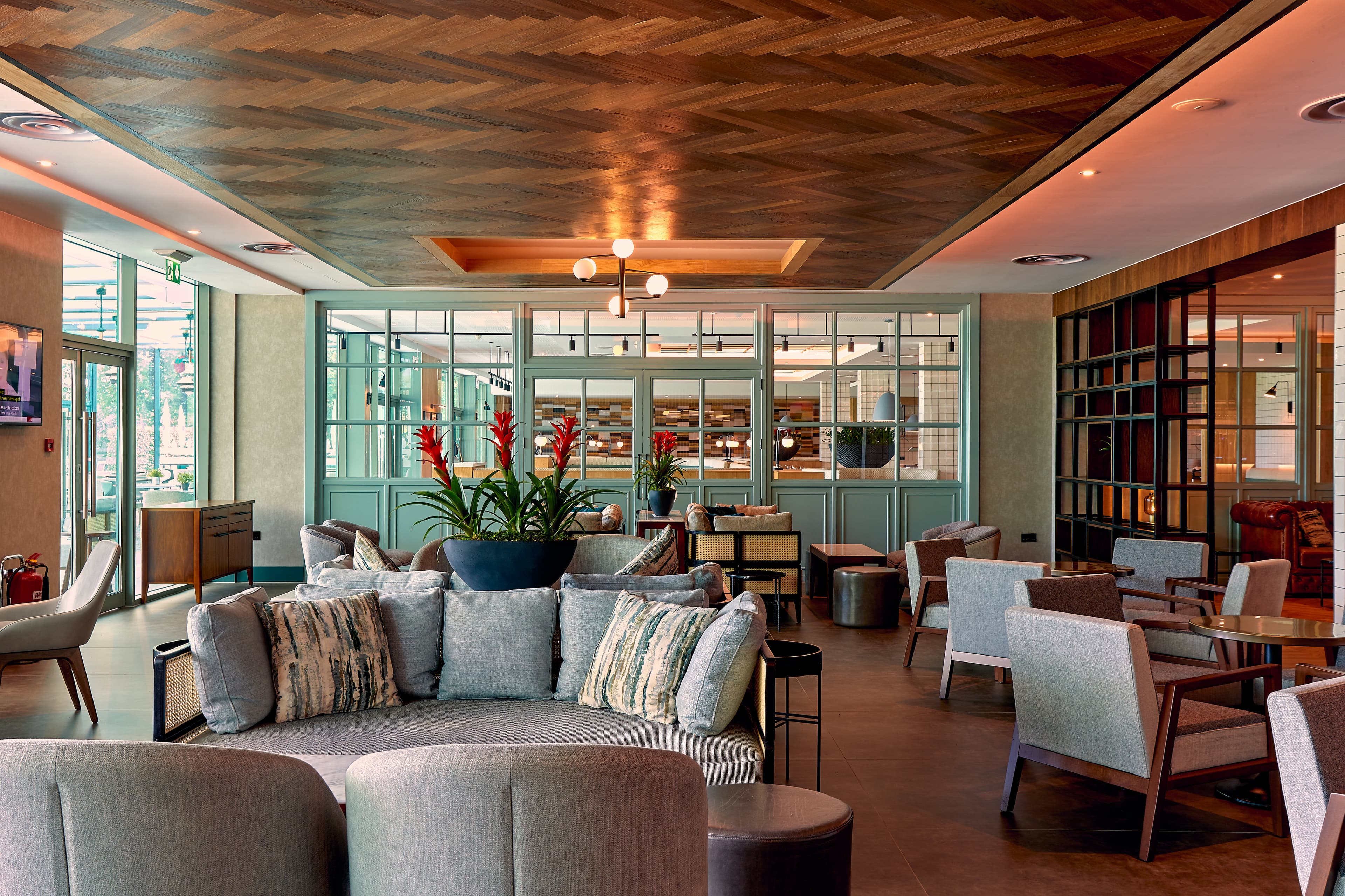 Hotel lounge area with grey sofas, armchairs, a herringbone wood ceiling and red tropical plants.