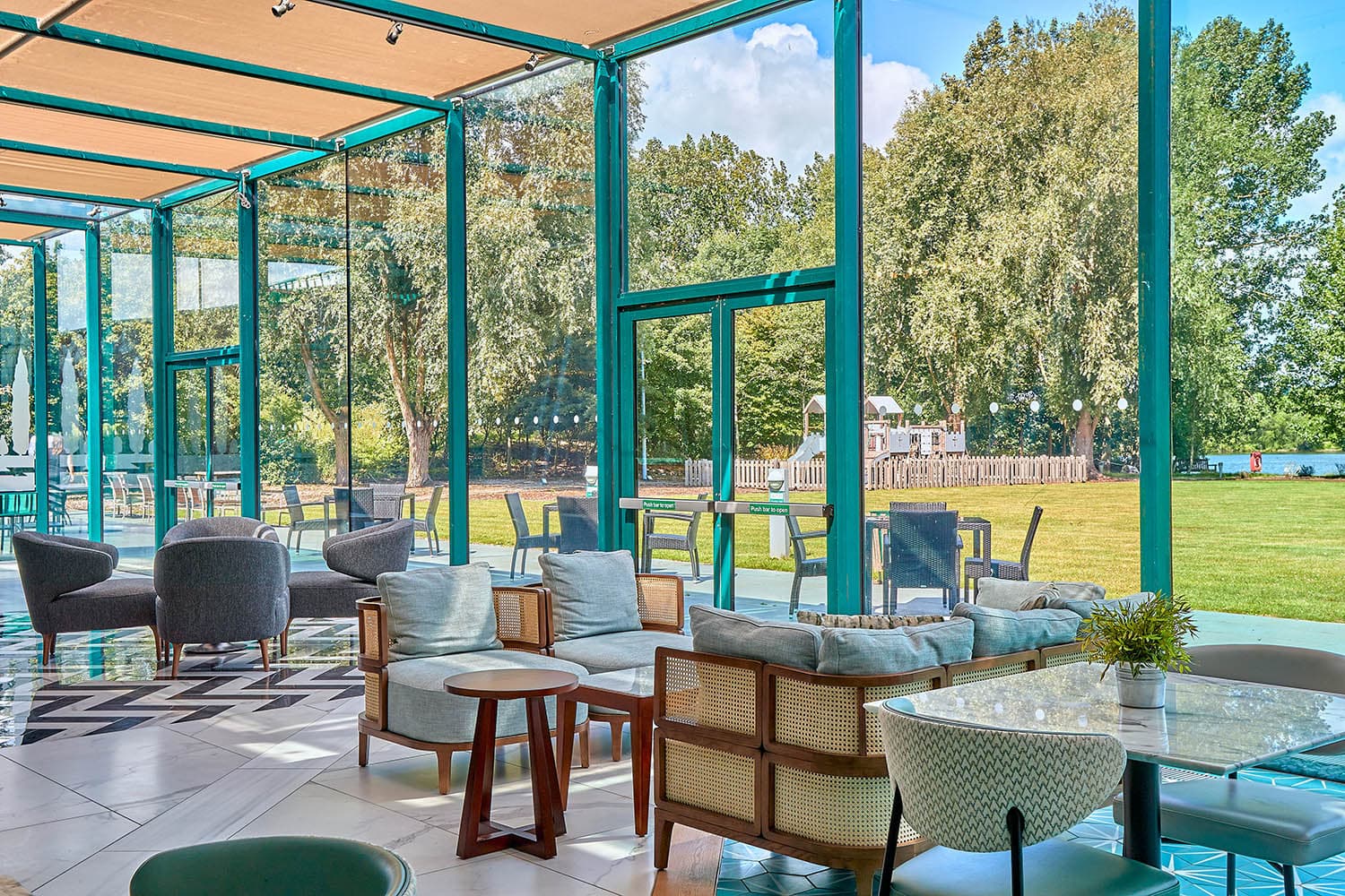 Light-filled conservatory lounge with rattan chairs overlooking a green lawn and children's play area.