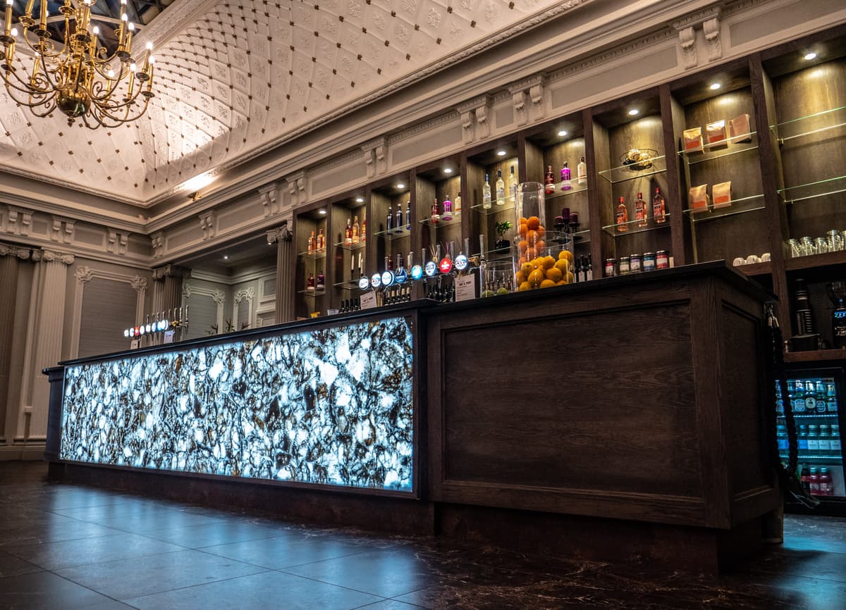Grand lounge bar counter with backlit stone front, draft taps and ornate plasterwork ceiling above.