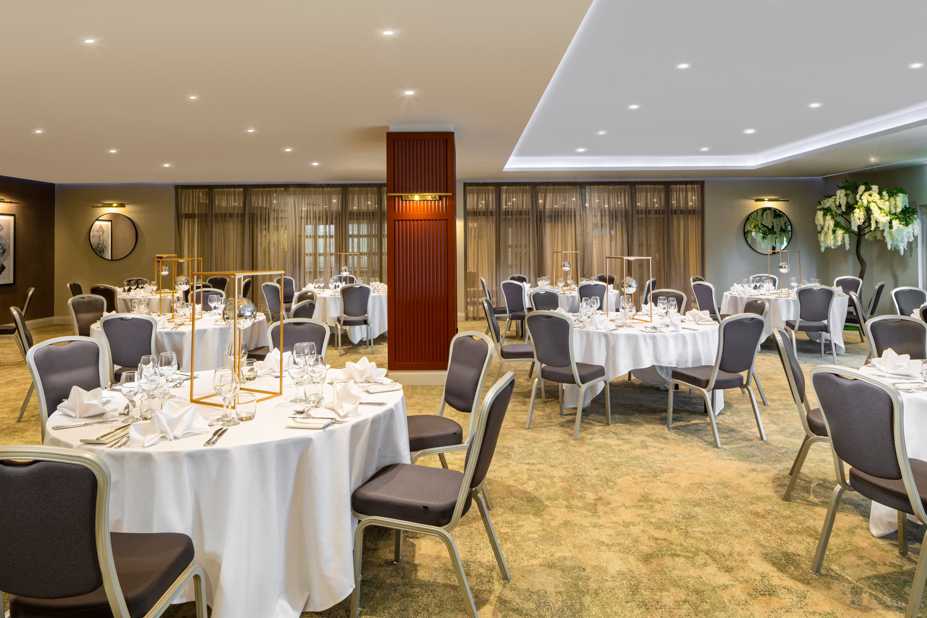 Banqueting room with round tables set with white linen, glassware and gold centrepieces.