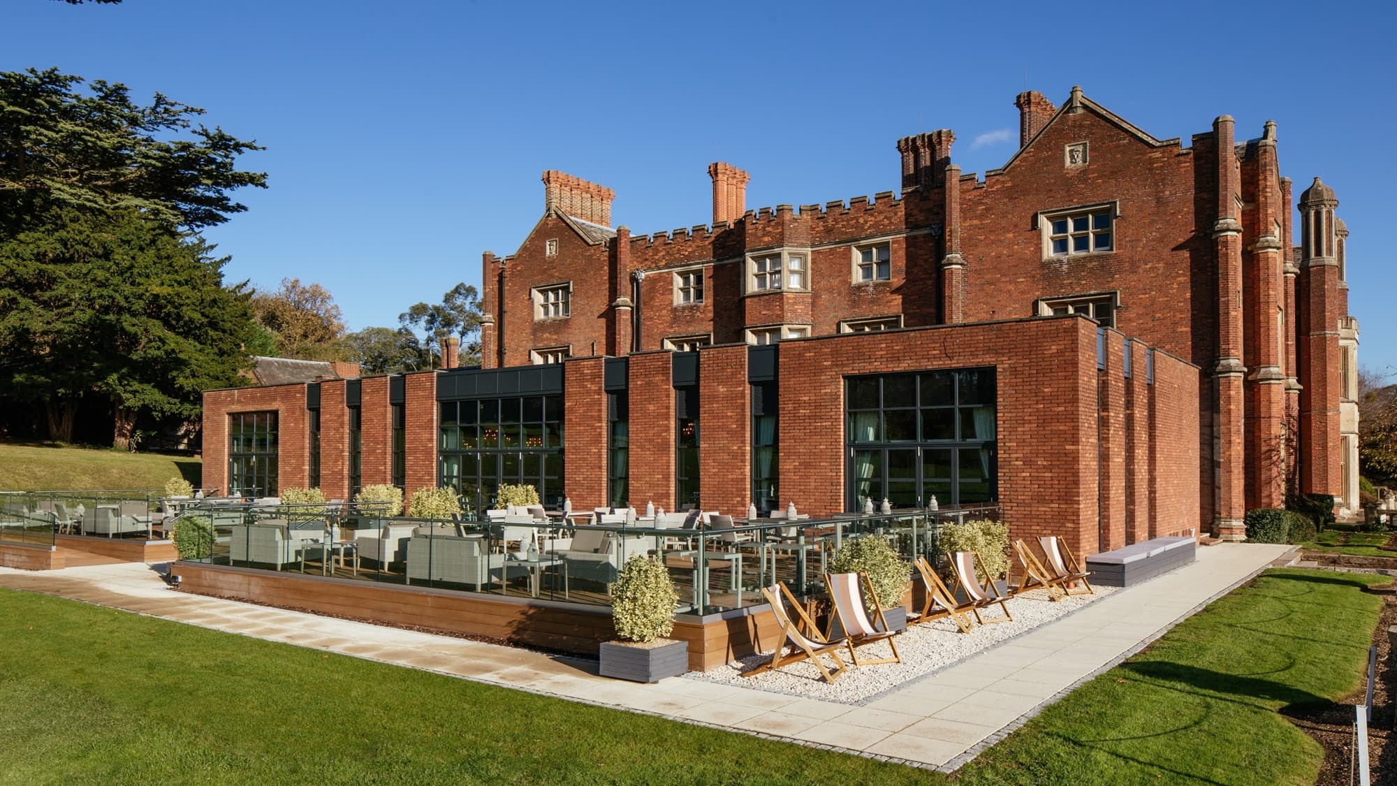 Red brick Tudor-style manor with a modern terrace, deckchairs and manicured lawns.