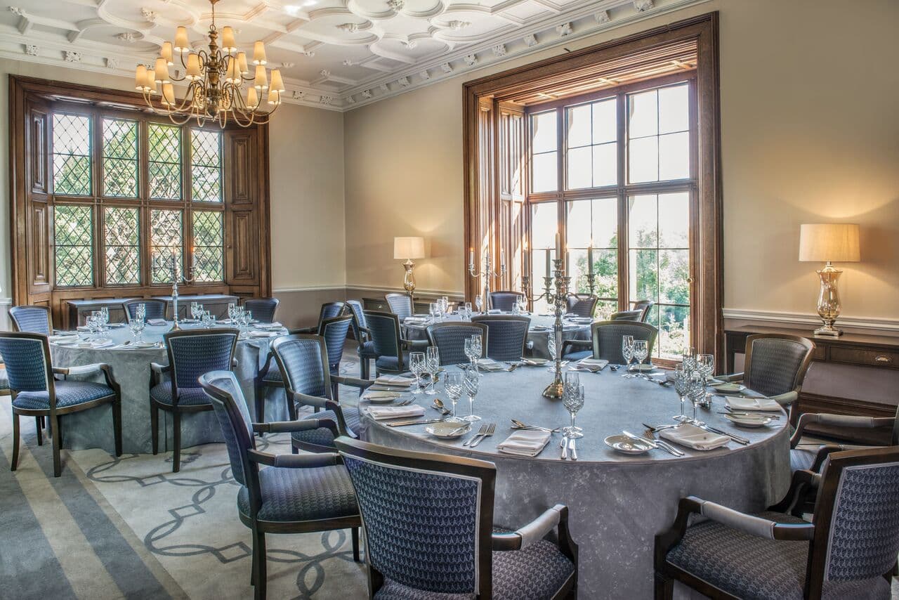 Formal dining room with round tables set for dinner and ornate plasterwork ceiling.