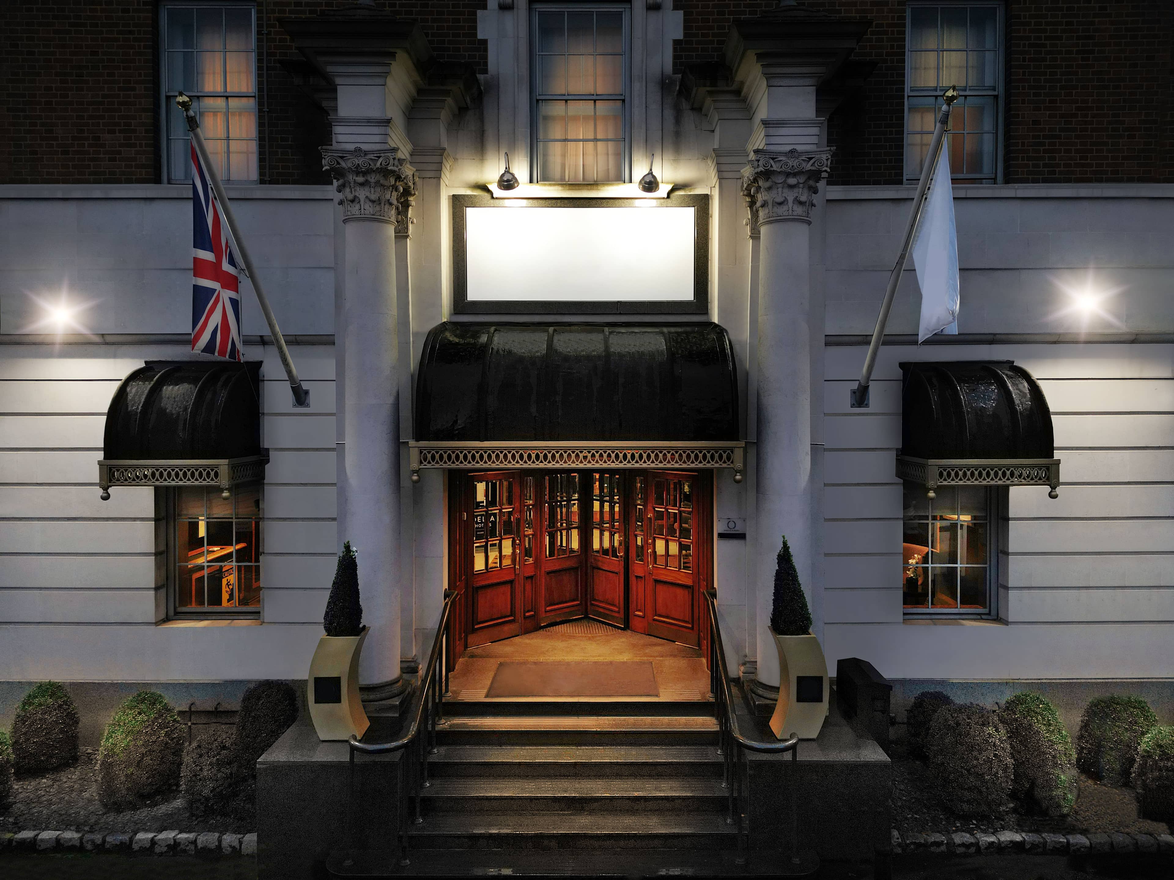 Floodlit hotel exterior at night with red doors, black canopy and Union Jack flag.