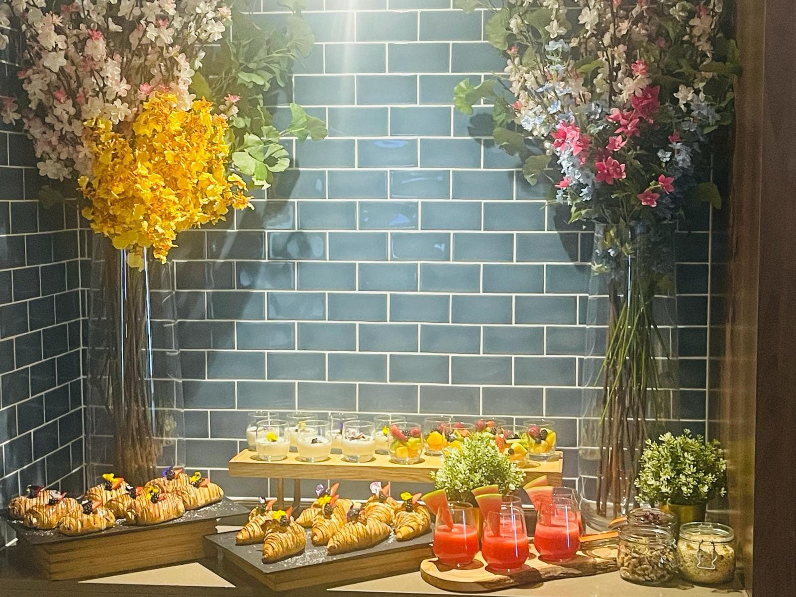 Buffet spread with croissants, fruit cups, pink drinks and tall floral arrangements against tiled wall.