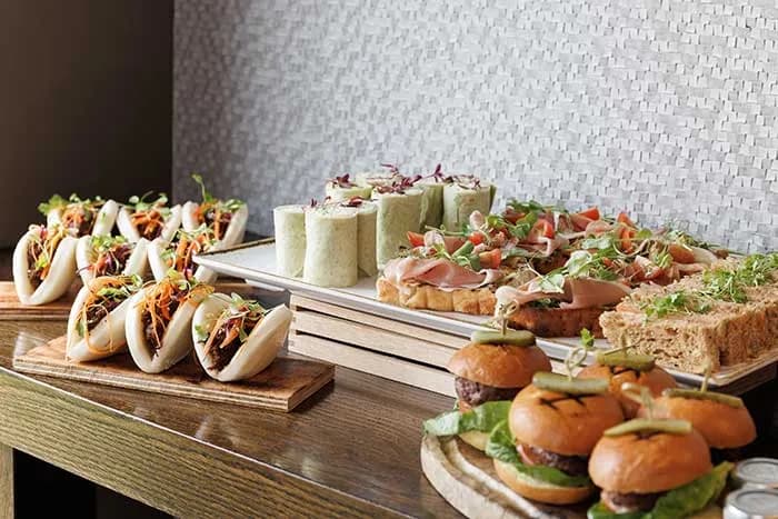 A buffet spread of bao buns, wraps, open sandwiches and mini burgers on wooden boards.