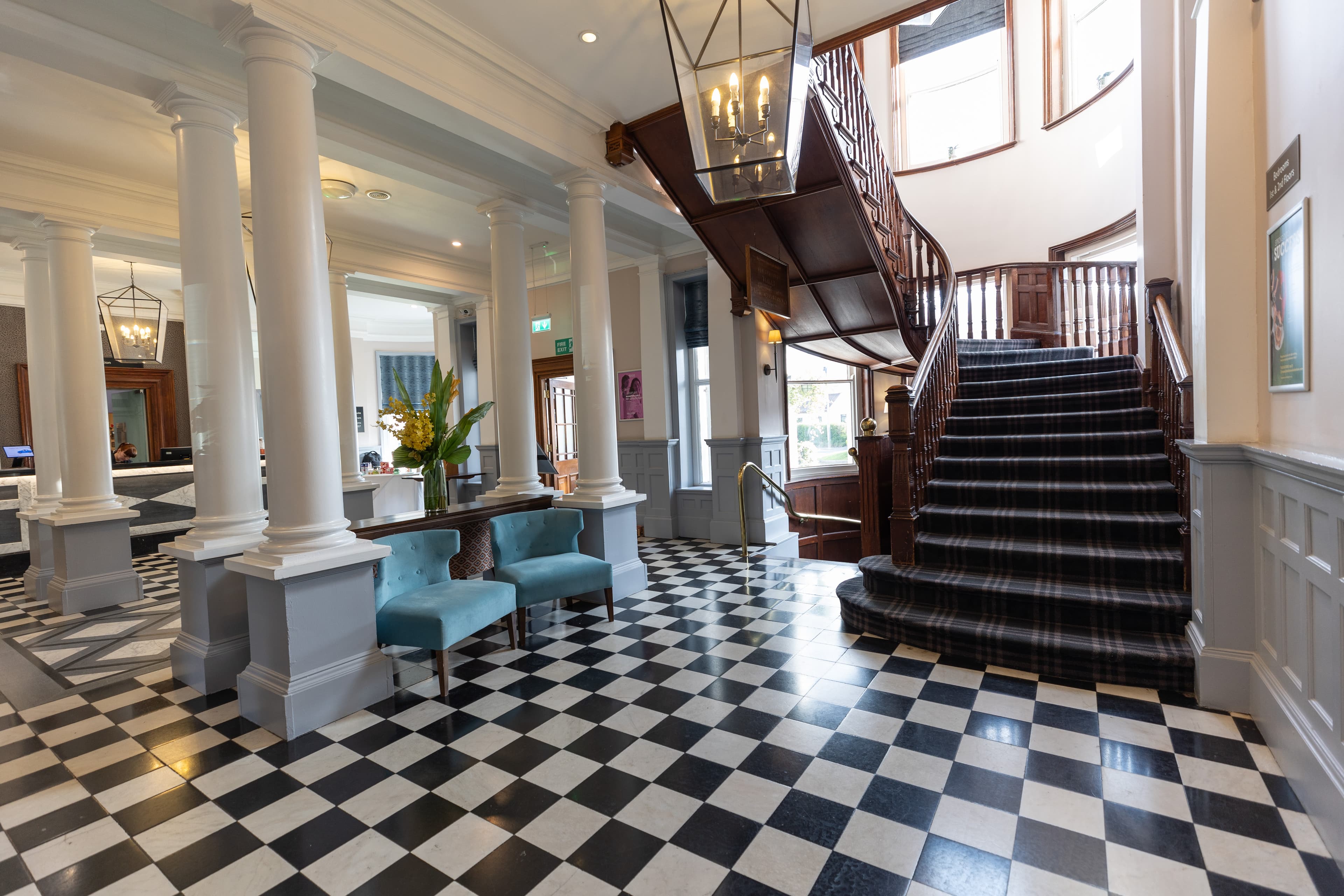 Hotel lobby with black and white tiled floor, white columns and a sweeping wooden staircase.