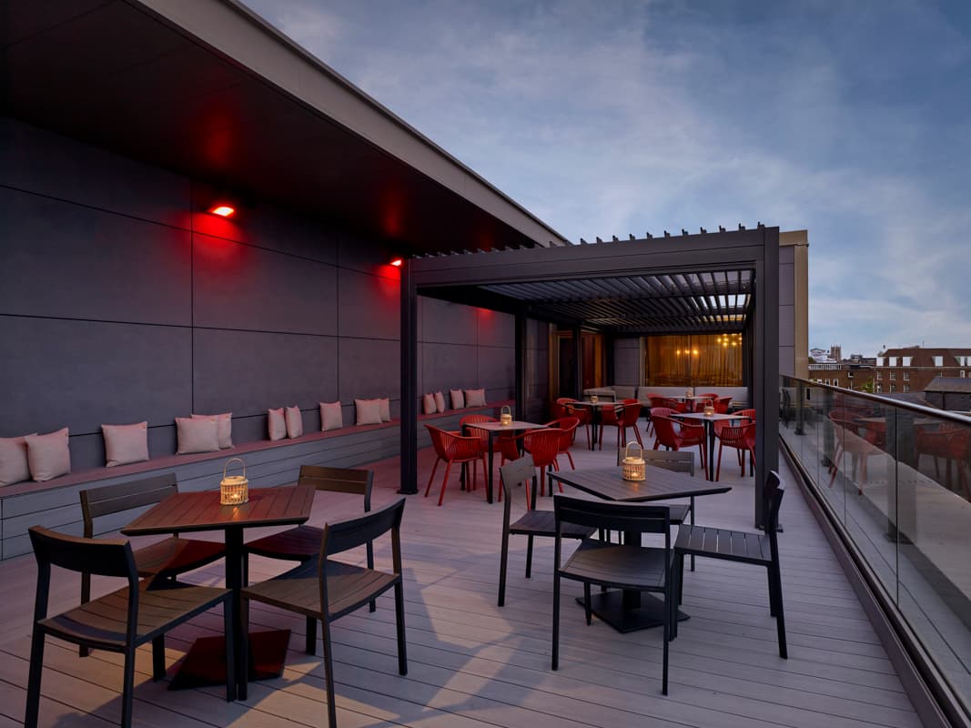 Rooftop terrace at dusk with red chairs, lantern-lit tables and a pergola.