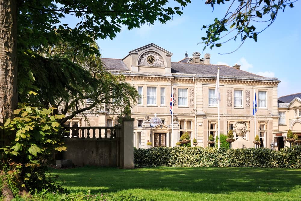 The stone exterior of Down Hall Hotel viewed through mature trees and lawns.