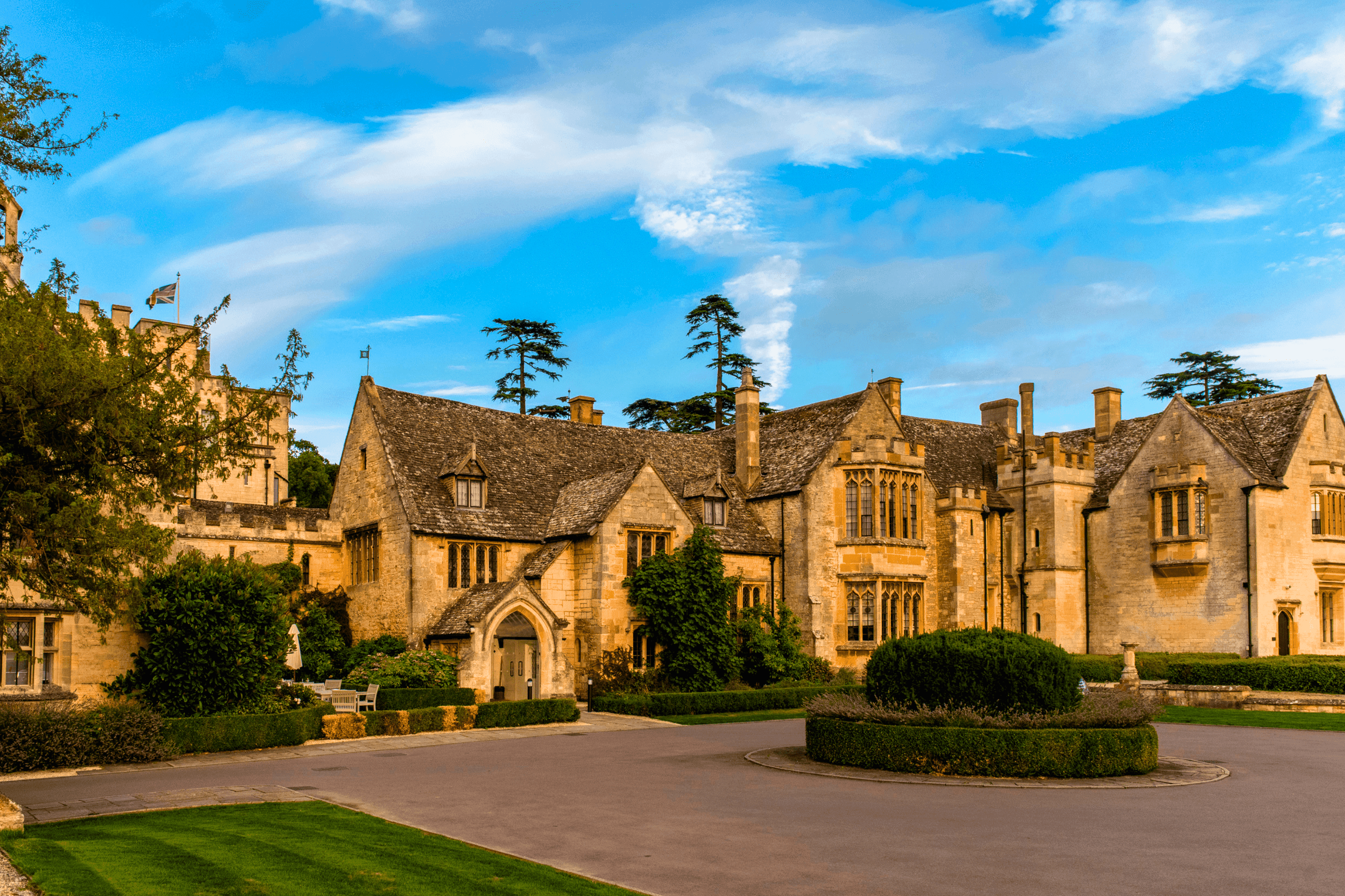 Honey-stone manor house exterior with circular driveway, topiary and mature cedar trees.