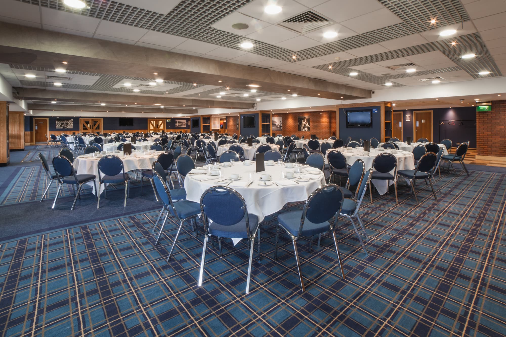 Large banqueting suite at Ewood Park with round tables, blue upholstered chairs and tartan carpet.