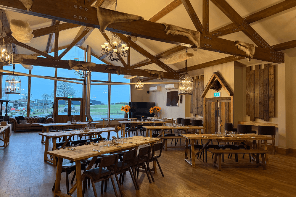 Rustic barn function room with wooden beams, long dining tables and large windows overlooking fields.