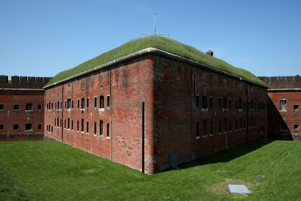 Exterior red brick rampart building at Fort Nelson with grass-covered roof and blue sky.