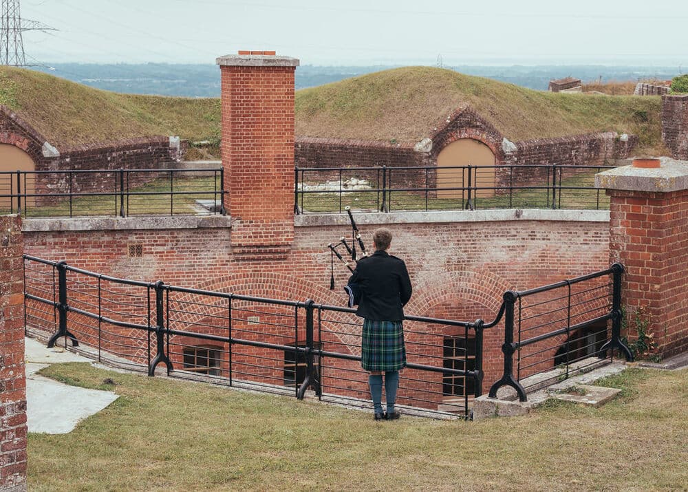 Bagpiper in a tartan kilt playing beside brick fortifications and grass ramparts.