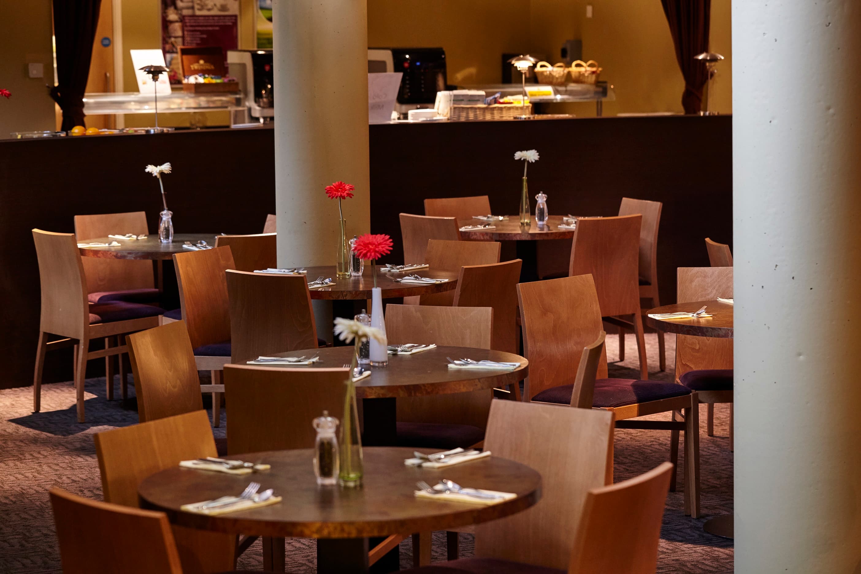 Restaurant dining area with round wooden tables, chairs and single stem flower arrangements.