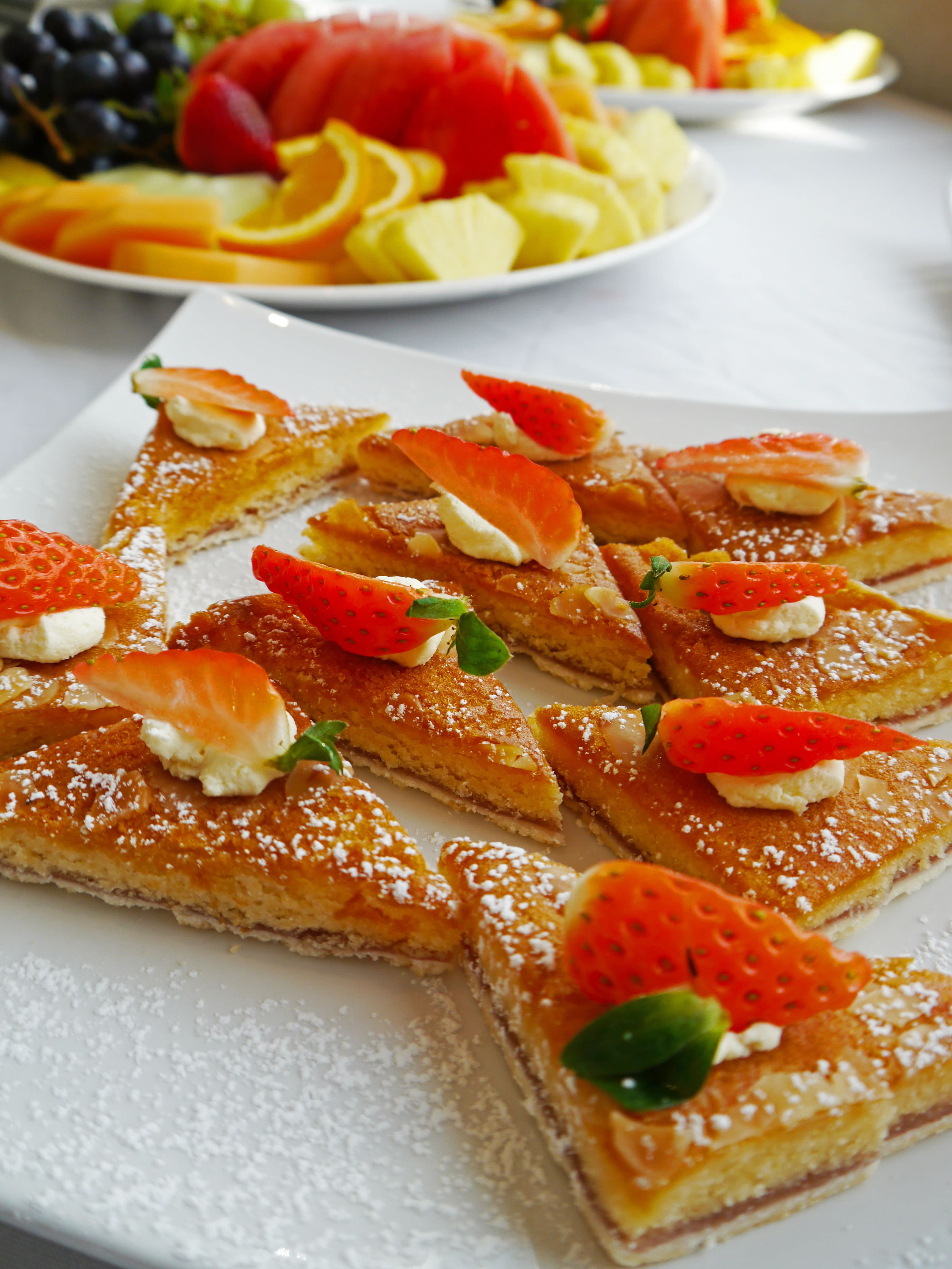 Triangular sponge cake slices topped with fresh strawberries and cream beside a fruit platter