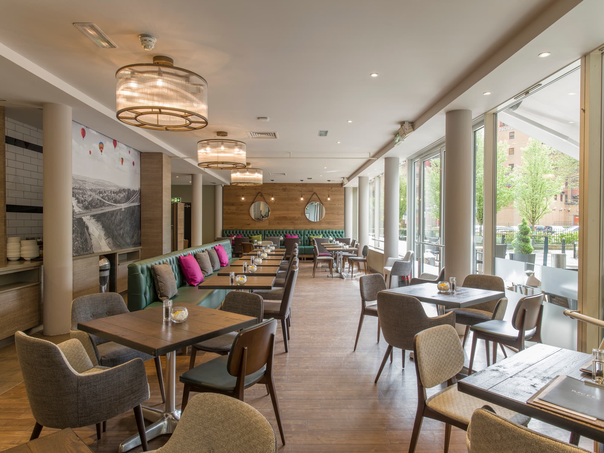 Bright hotel restaurant with green banquette seating, wooden tables and large street-facing windows.