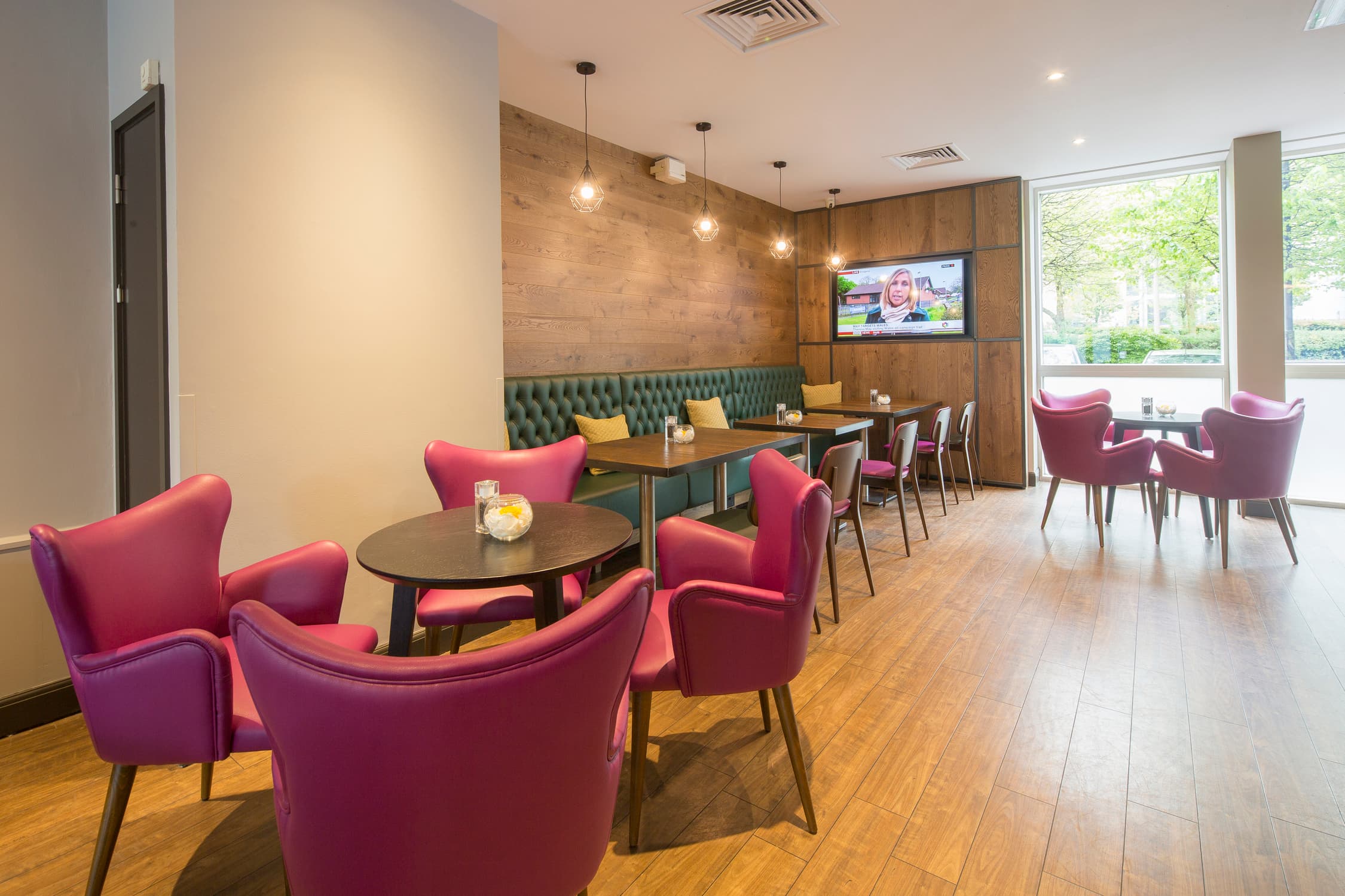Bright hotel lounge with pink armchairs, booth seating and a wall-mounted television.