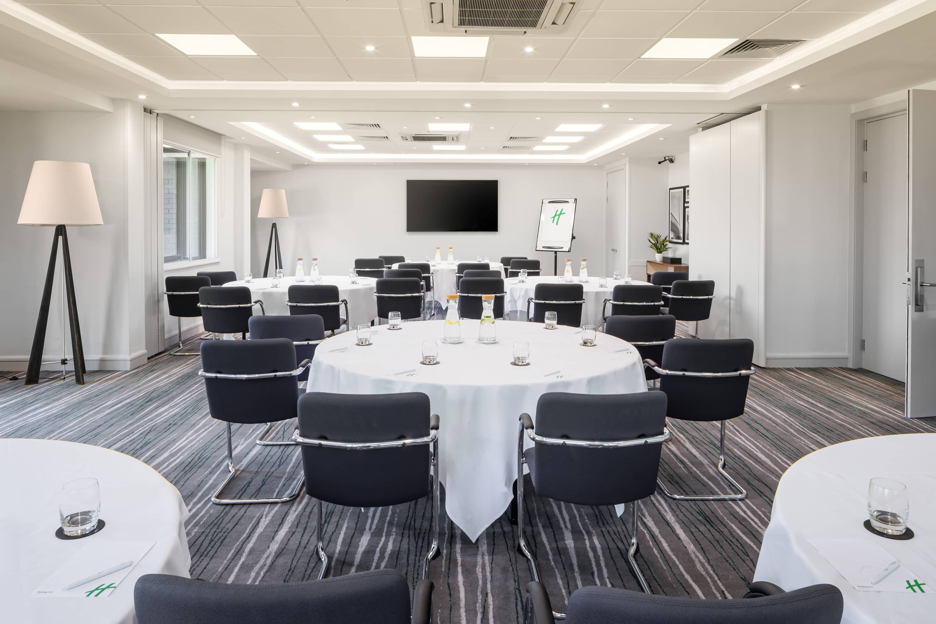 Conference room at Holiday Inn Aylesbury set with round tables, navy chairs and a wall-mounted screen.