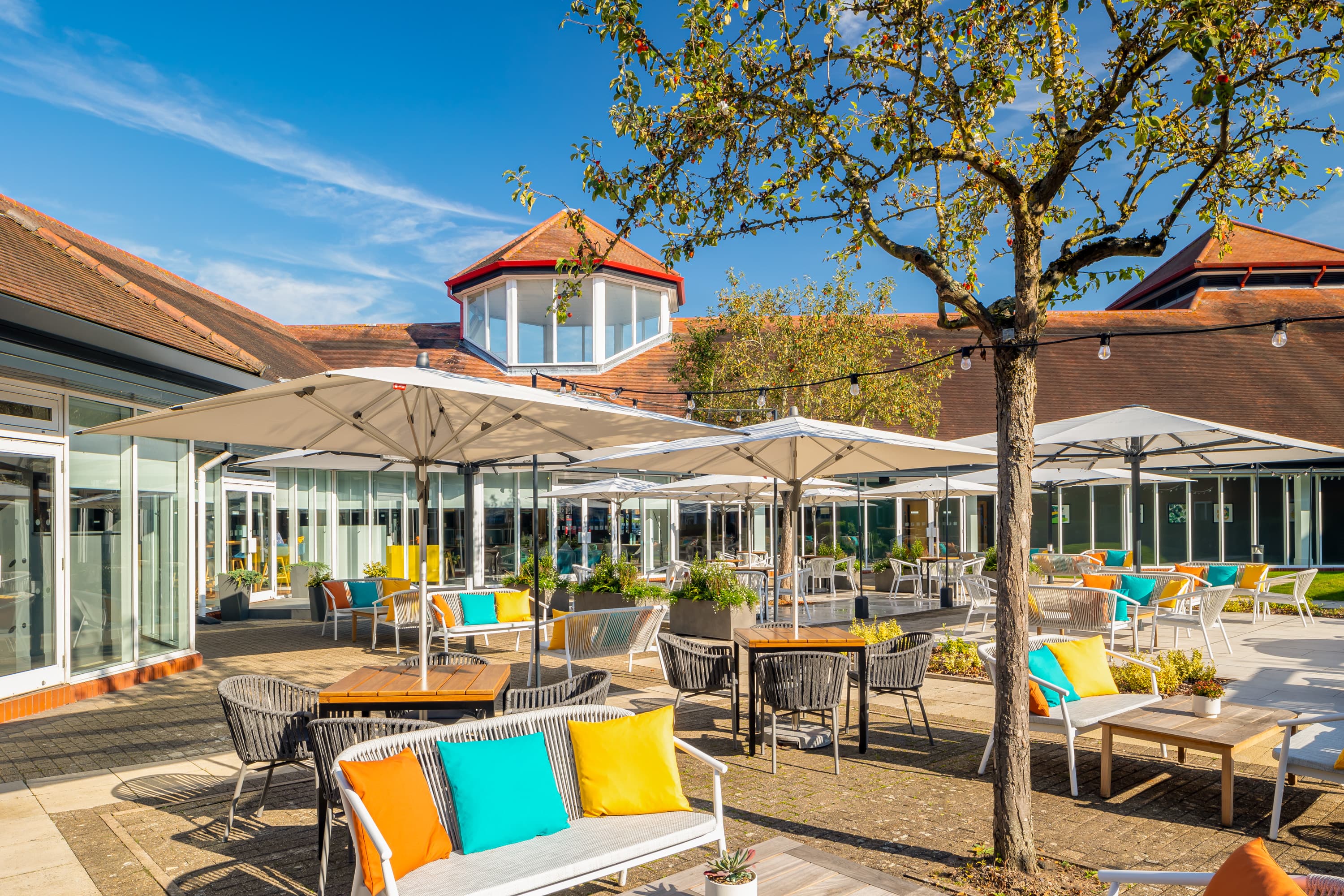 Sunny outdoor terrace with colourful cushioned seating, large parasols and festoon lighting.