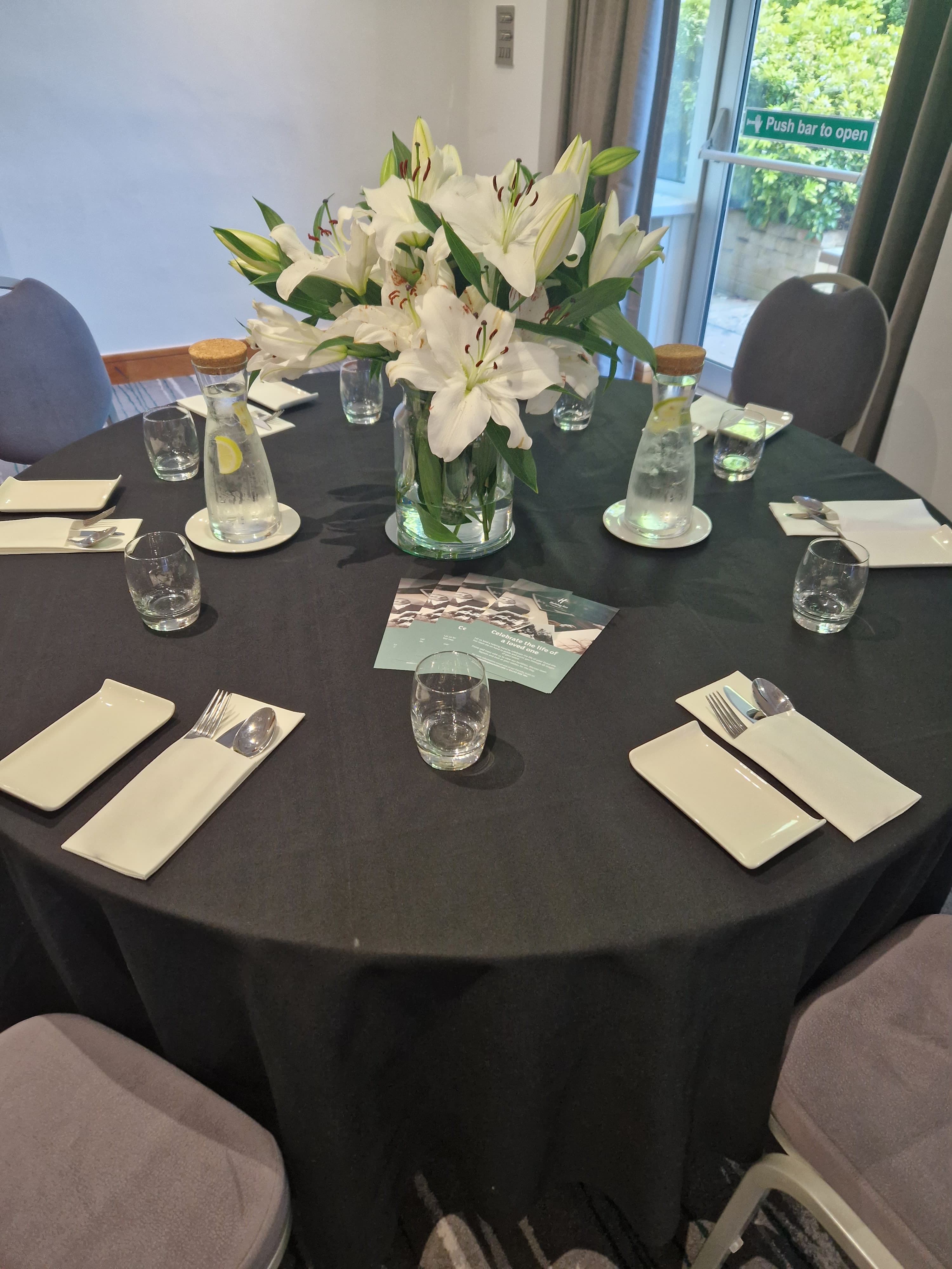 Round banquet table with white lily centrepiece, place settings and water carafes.