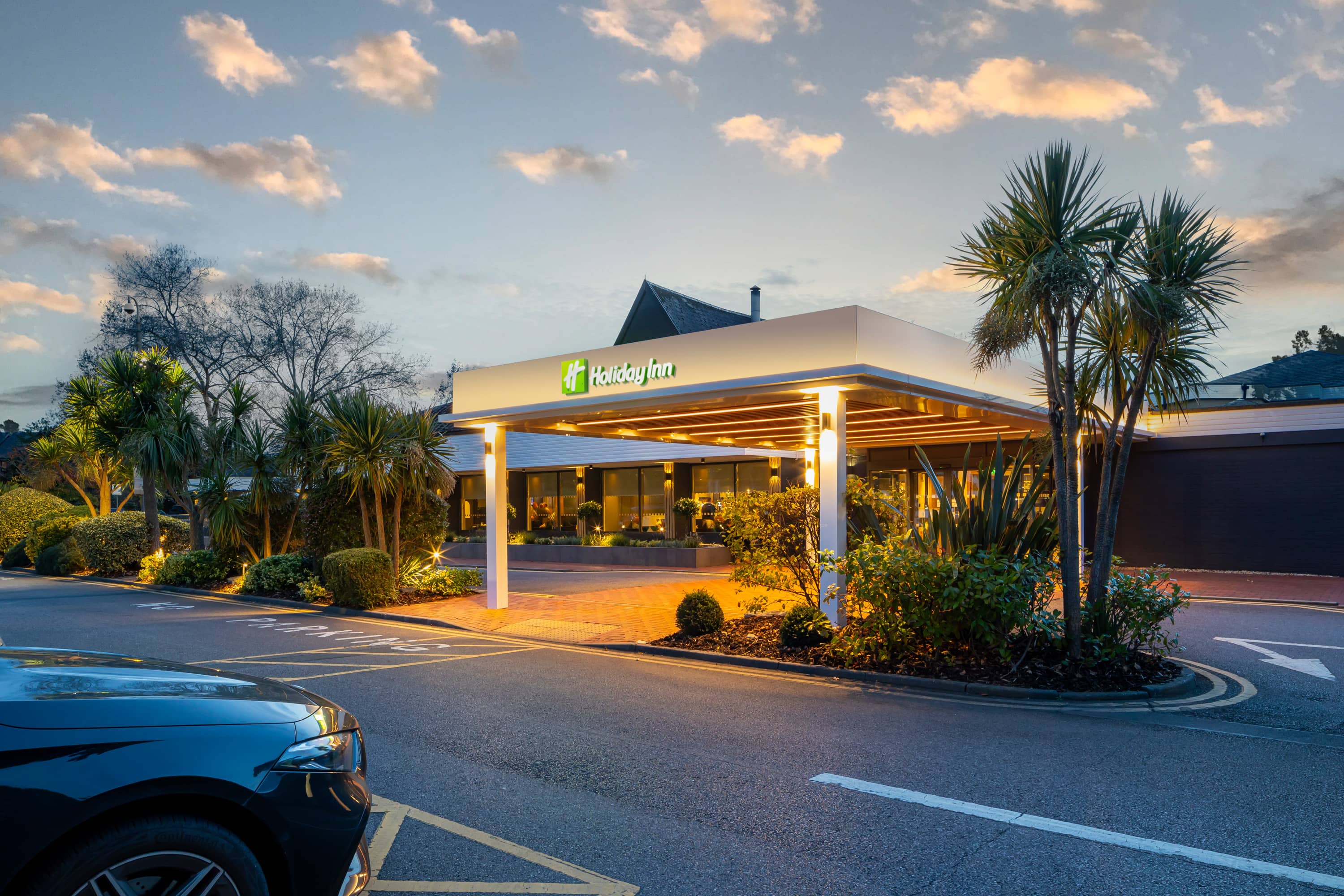 Illuminated Holiday Inn Reading South entrance canopy at dusk with palm trees and car park.