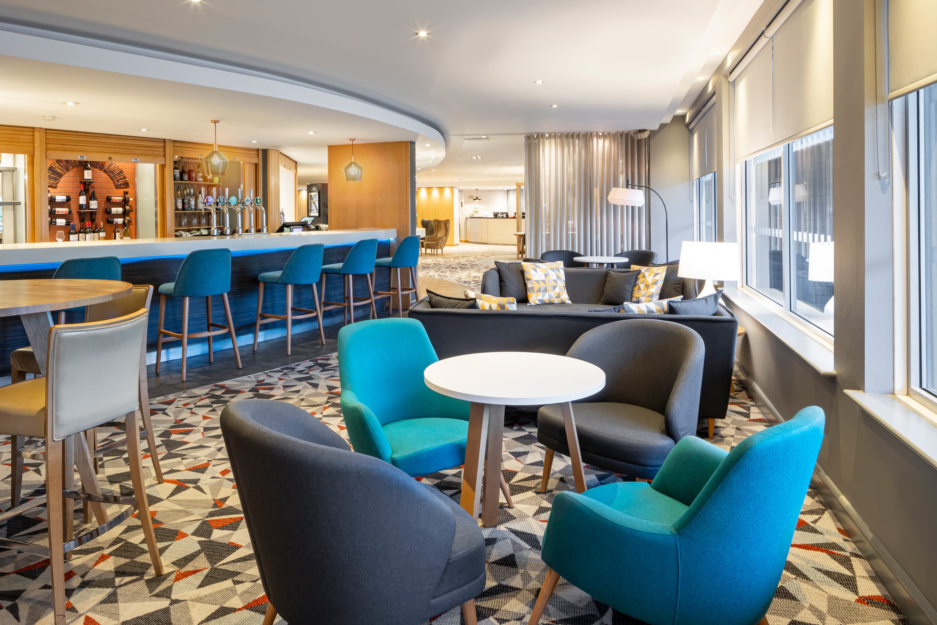 Hotel bar lounge with teal chairs, grey sofas and a long blue-lit counter.