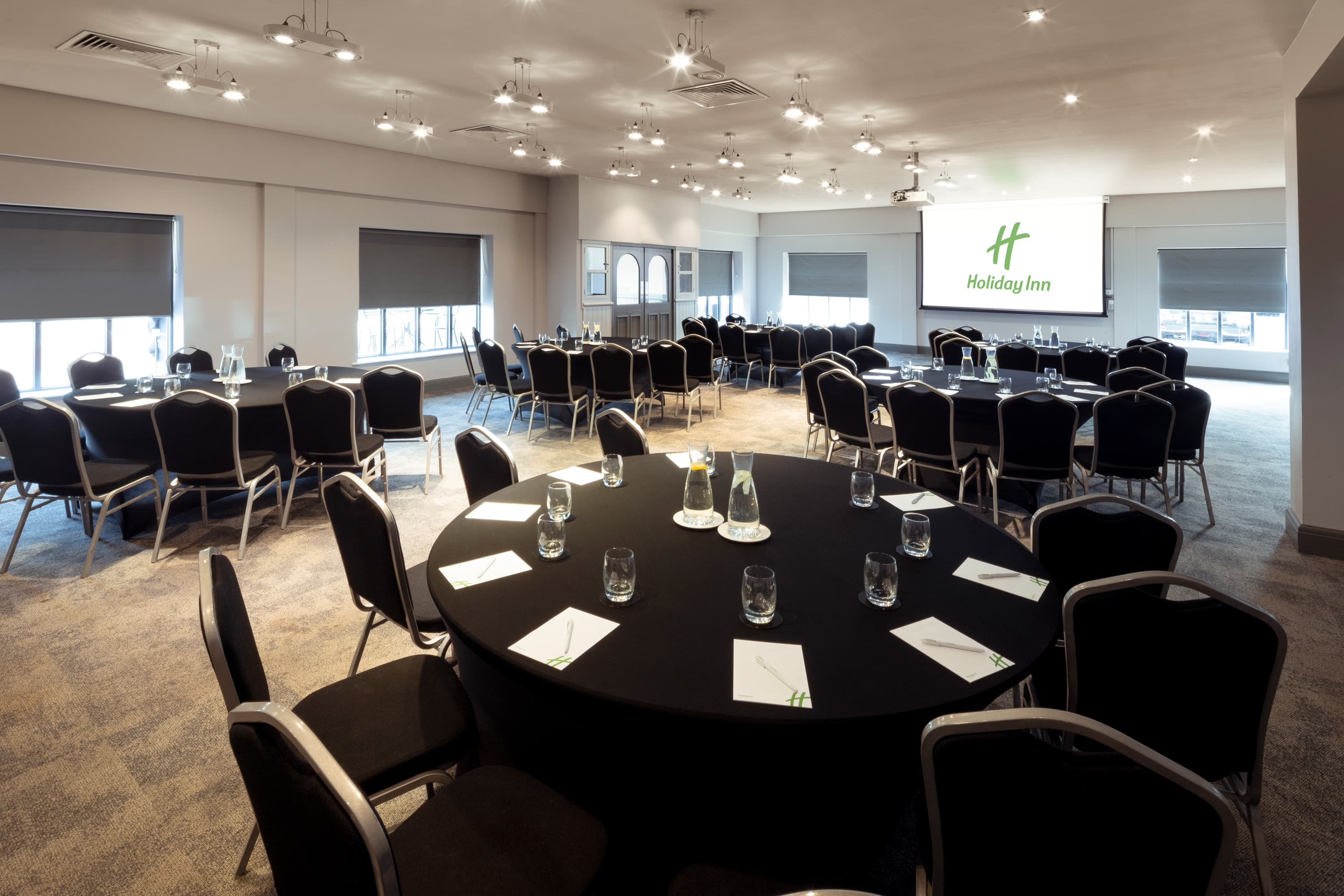 Large conference room with round black tables, banquet chairs and a projector screen.