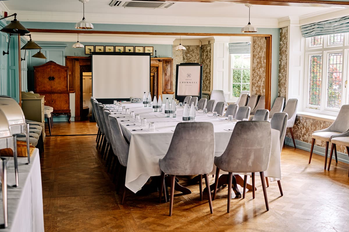 Boardroom-style meeting room with grey velvet chairs, white linen and a projection screen.