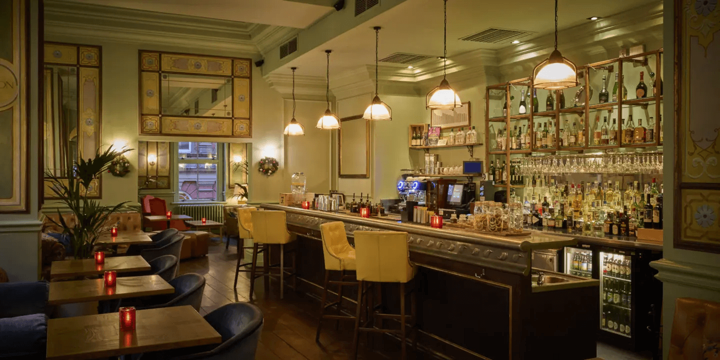 Warmly lit hotel bar with pale yellow stools, bottle shelves and candlelit seating area.
