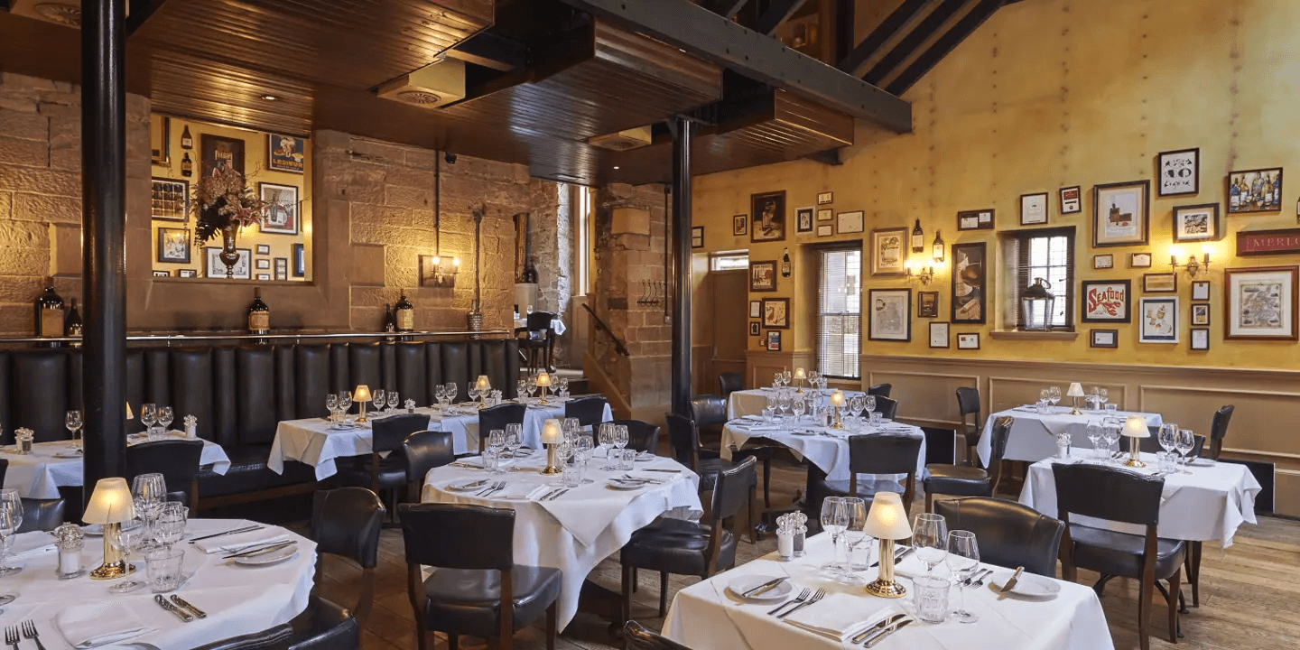 Restaurant dining room with white-clothed tables, leather chairs and framed artwork on yellow walls.